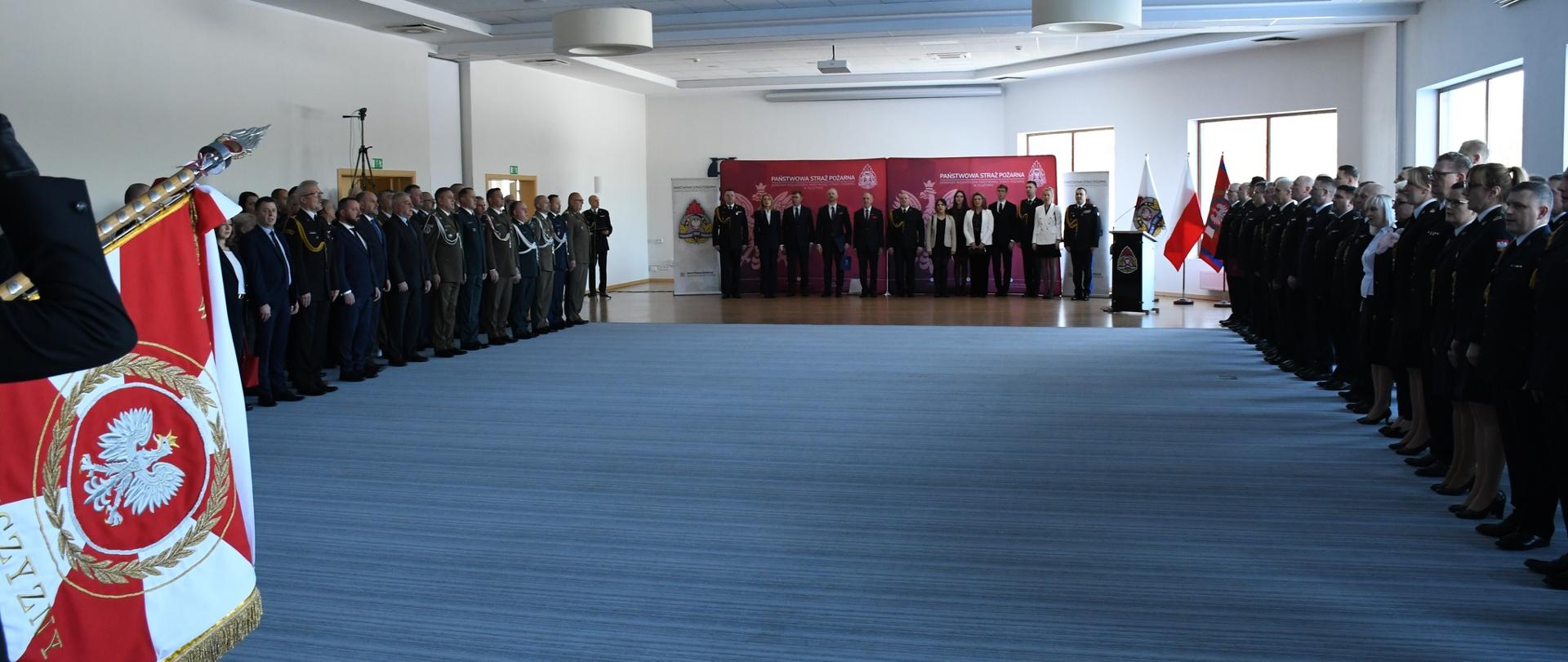 Duża sala. Z prawej, lewej i na wprost w oddali dużo zaproszonych gości. Zdanie obowiązków Warmińsko-Mazurskiego Komendanta Wojewódzkiego PSP
i Zastępcy Komendanta Wojewódzkiego PSP
