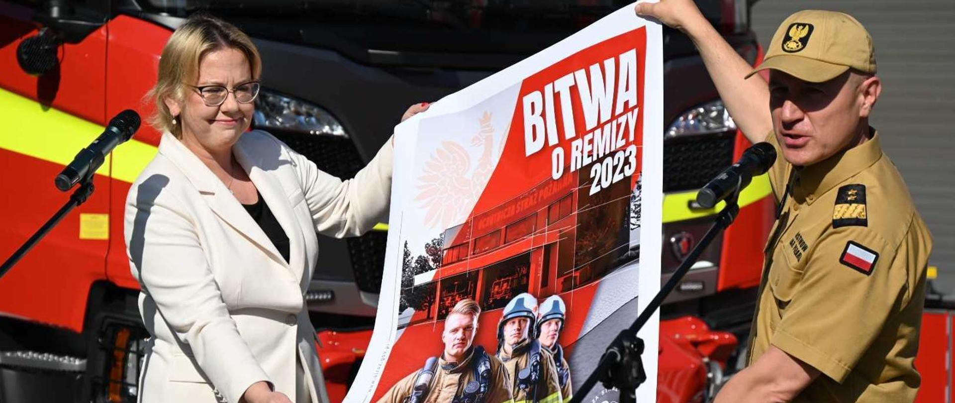 Kobieta w białej marynarce i umundurowany strażak prezentują plakat z napisem "Bitwa o remizy 2023"