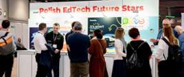 Widok na białe stoisko z napisem Polish EdTech Future Stars, przed stoiskiem grupa oglądających ludzi, w tle duża hala.