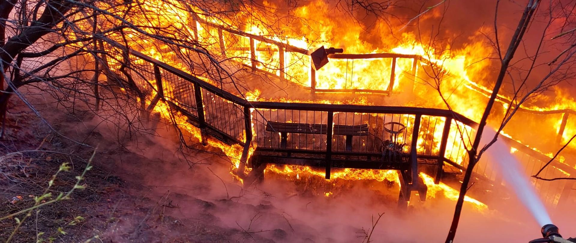 Zdjęciu ukazuje schody, które są całe w płomieniach. W dolnym rogu widać prądownicę wodną, z której wylatuje woda.