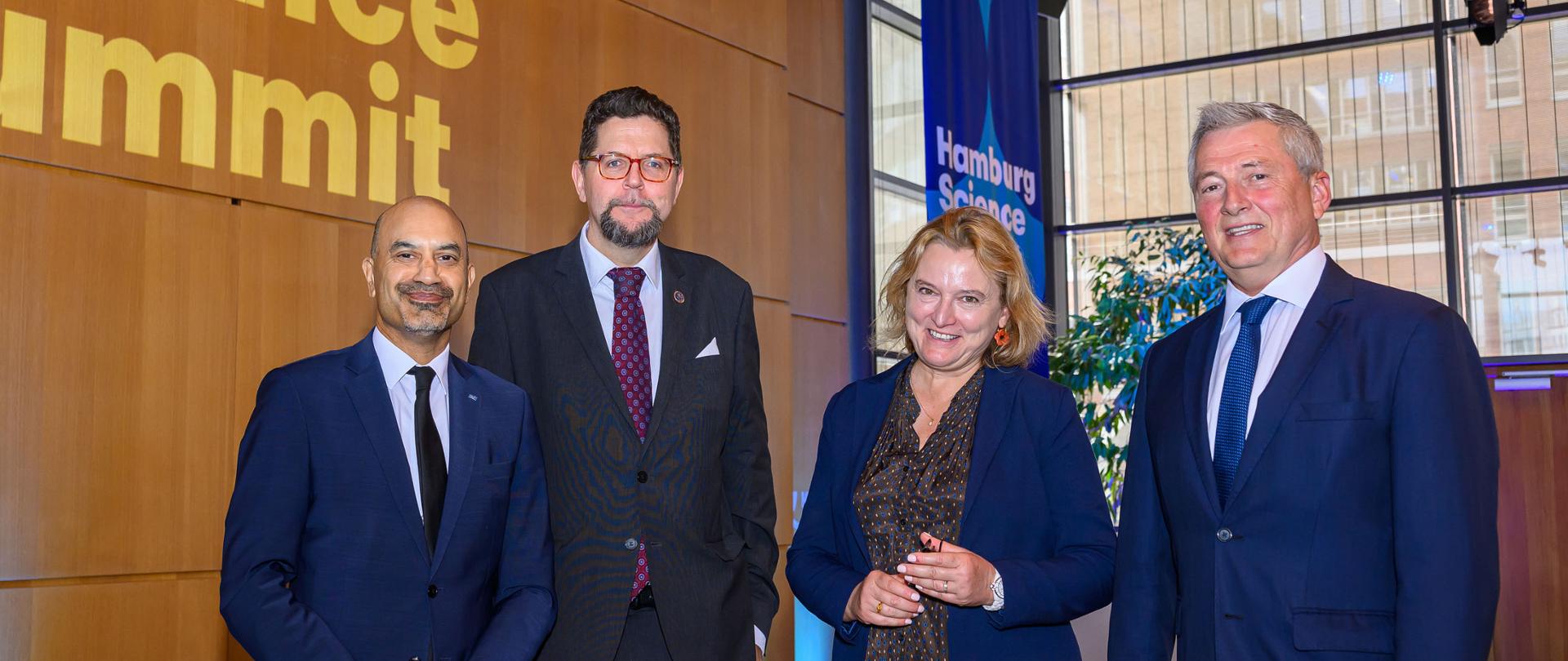 Pod brązową ścianą z napisem Hamburg Science Summit stoją wiceminister Szeptycki, kobieta w niebieskiej bluzce i dwóch mężczyzn w garniturach, w tle okno.