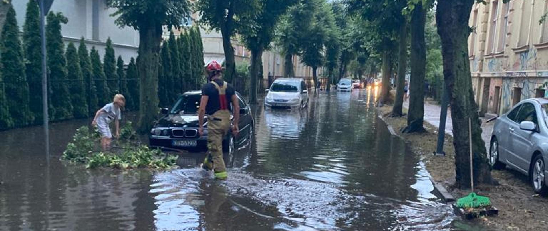 ZAlana ulica a na poboczu stojące zaparkowane samochody Po środku strażak udrażniający studzienkę odpływowa 