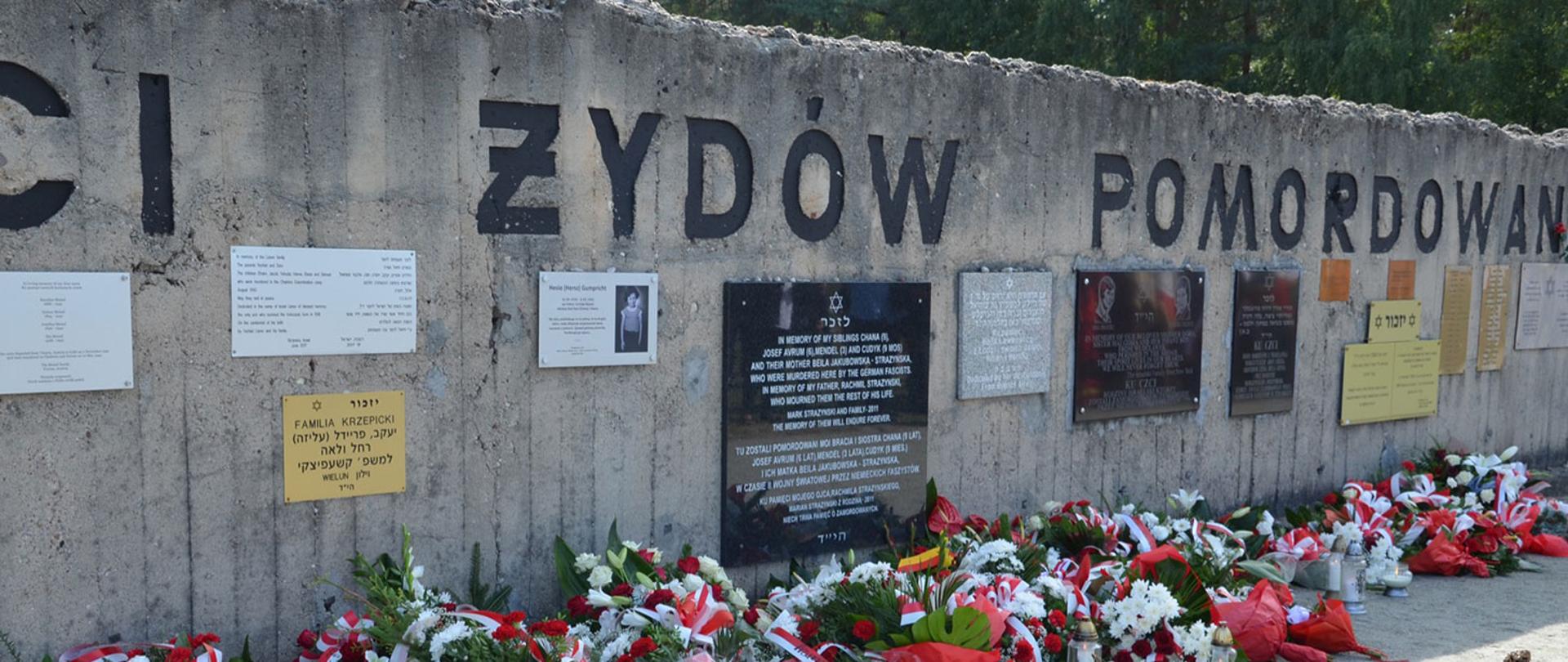 Zdjęcie przedstawia mur, z tablicami nieupamiętniającymi zagładę pomordowanych ludzi w obozie. Pod murem złożone kwiaty.