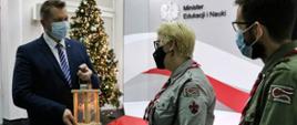 Minister Edukacji i Nauki Przemysław Czarnek trzymający Betlejemskie Światło Pokoju, obok stoją harcerze, a, w tle udekorowana choinka.