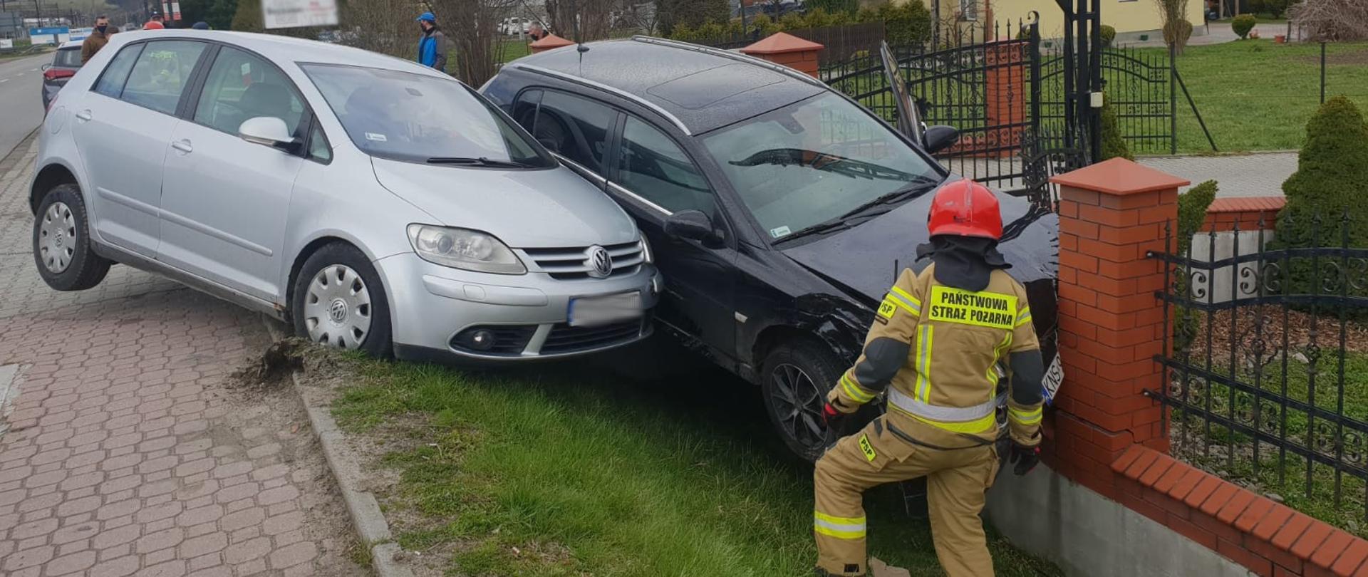 Dwa samochody osobowe jeden jasnego koloru a drugi ciemnego koloru poza drogą obok siebie. Ciemny uderzył w słupek ogrodzenia. Przed samochodami stoi strażak. W tle budynki oraz tablice ogłoszeniowe.