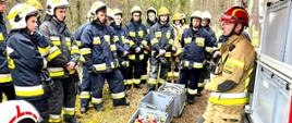 Po stronie lewej widzimy kursantów, którzy słuchają instruktora znajdującego się po prawej stronie. Na środku sprzęt używany podczas pożarów lasu. Widać również część przyczepy gaśniczej. W tle widać las. 