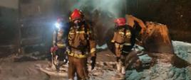 Zdjęcie przedstawia strażaków podczas gaszenia palącej się ładowarki kołowej. Strażacy zabezpieczeni w sprzęt ochrony dróg układu oddechowego. Na zdjęciu widać ładowarkę, która uległa spaleniu oraz pianę gaśniczą. 
