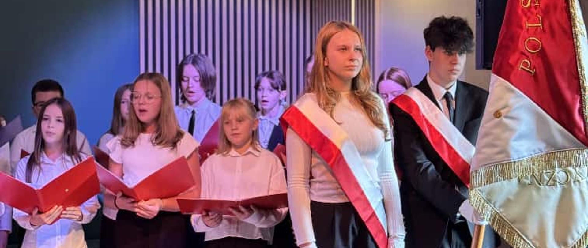 Na zdjęciu widać osoby śpiewające w chórze i poczet sztandarowy szkoły muzycznej. W tle widać zieloną ścianę oraz drewniane lamele na ścianie