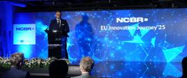 Minister Marcin Kulasek stoi przy mównicy na tle niebieskiej ściany z logo NCBR