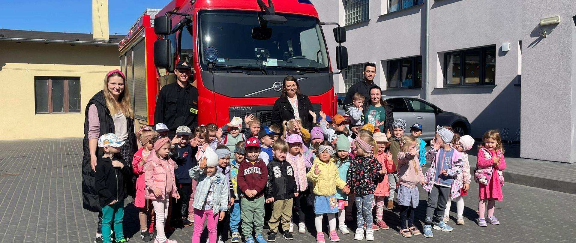 Zdjęcie grupowe przedstawiające dzieci uczestniczące w prewencji społecznej realizowanej w placówce dydaktycznej na tle samochodu pożarniczego,. Dzieci wspólnie ze strażakami realizującymi pokaz sprzętu stoją na palcu przy przedszkolu., 