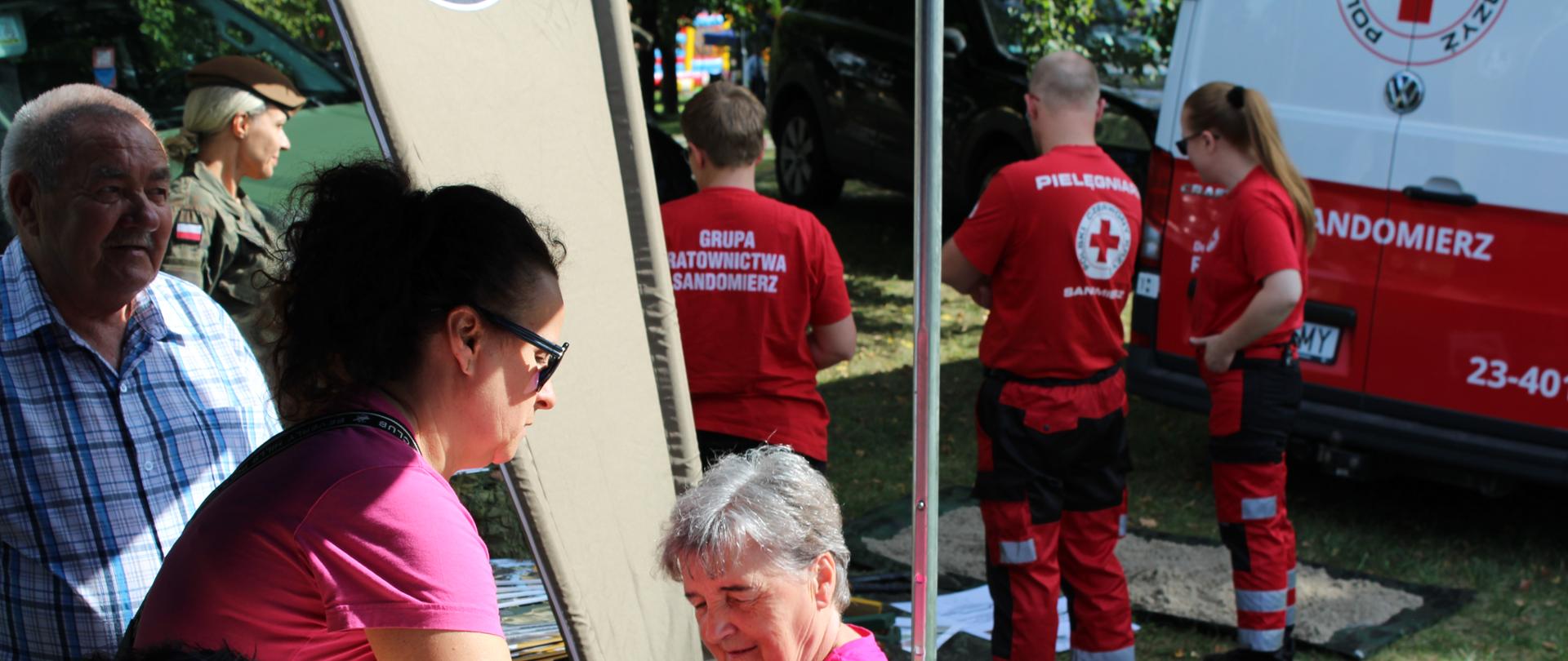 Na głównym planie przedstawicielka WSSE w różowej bluzce , wykonująca pomiar ciśnienia krwi pani, która siedzi na krześle. Po lewej stronie pan towarzyszący żonie. Pani ubrana w różową bluzkę. W tle grupa ratownictwa ubrana w czerwone koszulki