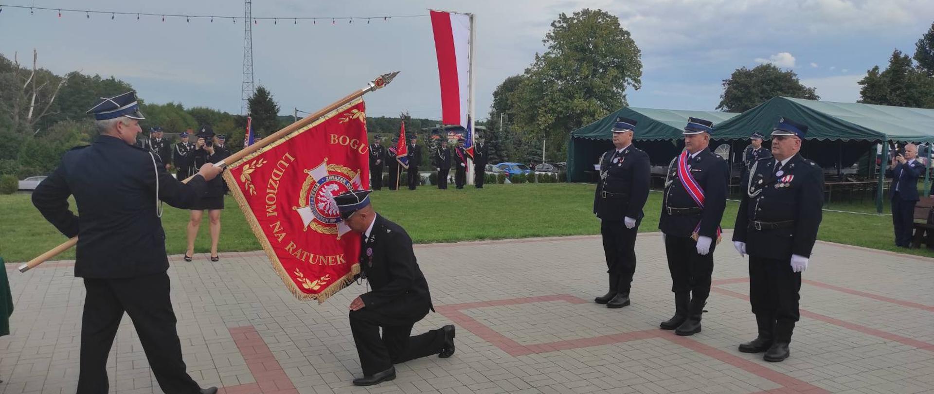 Na zdjęciu klękający strażak OSP całuje sztandar, za nim stoi poczet sztandarowy. W tle widać namioty, flagę Polski oraz stojące poczty sztandarowe.