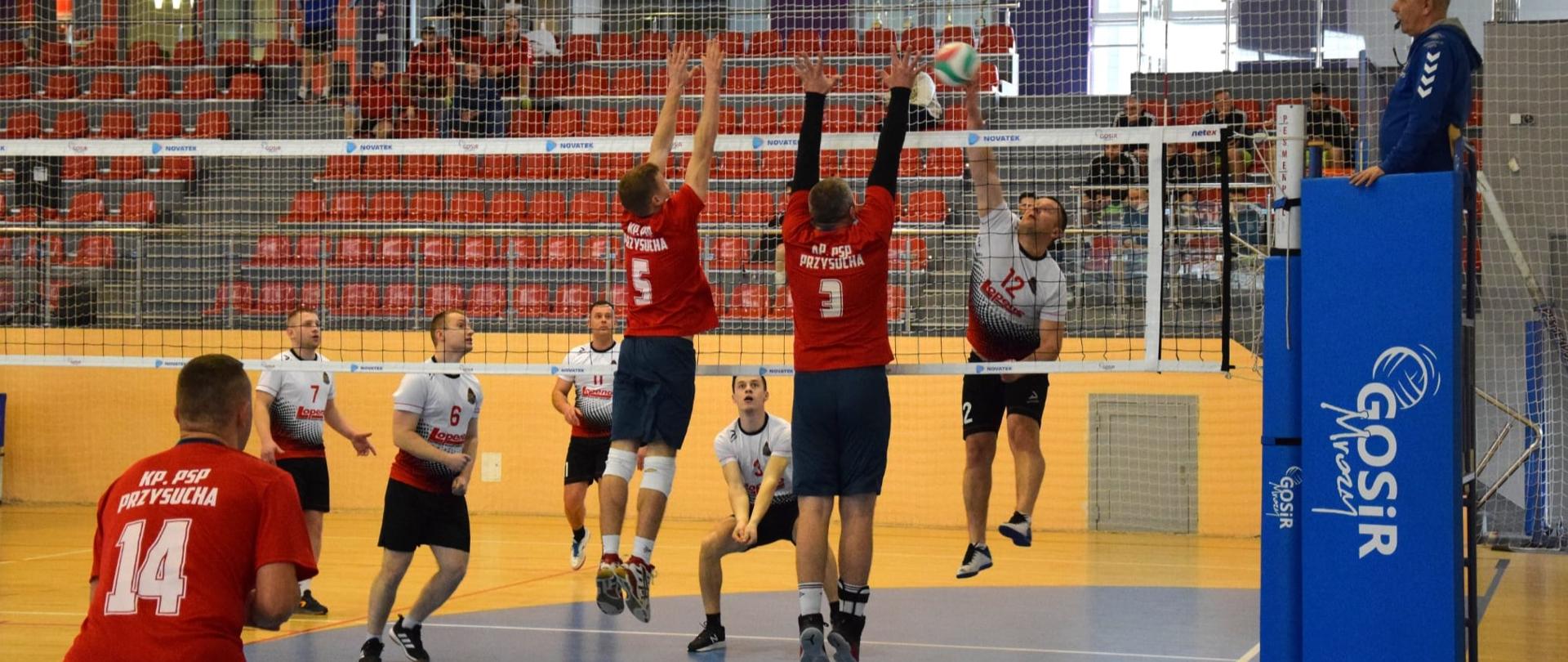 Poziome zdjęcie przedstawia moment meczu siatkówki na hali sportowej. Skupia się na akcji przy siatce, gdzie zawodnicy walczą o piłkę.
W centralnej części kadru trzech zawodników znajduje się w powietrzu, skacząc do bloku lub ataku. Dwóch z nich, ubranych w czerwone koszulki z numerami "5" i "3" i napisem "KP PSP PRZYSYCHA", są wyraźnie widoczni, z rękami wyciągniętymi nad siatką. Trzeci zawodnik, w białej koszulce z czarnymi wstawkami i numerem "12", również skacze do piłki, która jest widoczna nad jego rękami. Dwóch zawodników z drużyny Komendy Powiatowej Państwowej Straży Pożarnej w Przysusze blokuje atak zawodnika z drużyny przeciwnej.
Na boisku widoczni są także inni zawodnicy obu drużyn. Jeden z zawodników w czerwonej koszulce z numerem "14" i napisem "KP PSP PRZYSUCHA" stoi na pierwszym planie po lewej stronie.
Siatka do siatkówki jest rozciągnięta przez środek kadru. Po prawej stronie siatki stoi słupek z niebieską osłoną, na której widnieje logo "GOSiR Mrozy". Obok słupka stoi sędzia, patrzący na boisko.
W tle widać trybuny z pustymi czerwonymi krzesełkami oraz górne partie hali sportowej z widownią i oświetleniem. Podłoga boiska jest w kolorze niebieskim i brązowym z białymi liniami. Zdjęcie oddaje dynamikę i napięcie meczu siatkówki.
