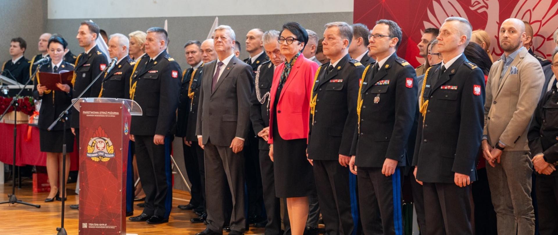 Uczestnicy uroczystości stoją w szeregu na sali, w tym oficerowie PSP w galowych mundurach oraz zaproszeni goście. Na pierwszym planie znajduje się podium z emblematem Państwowej Straży Pożarnej.