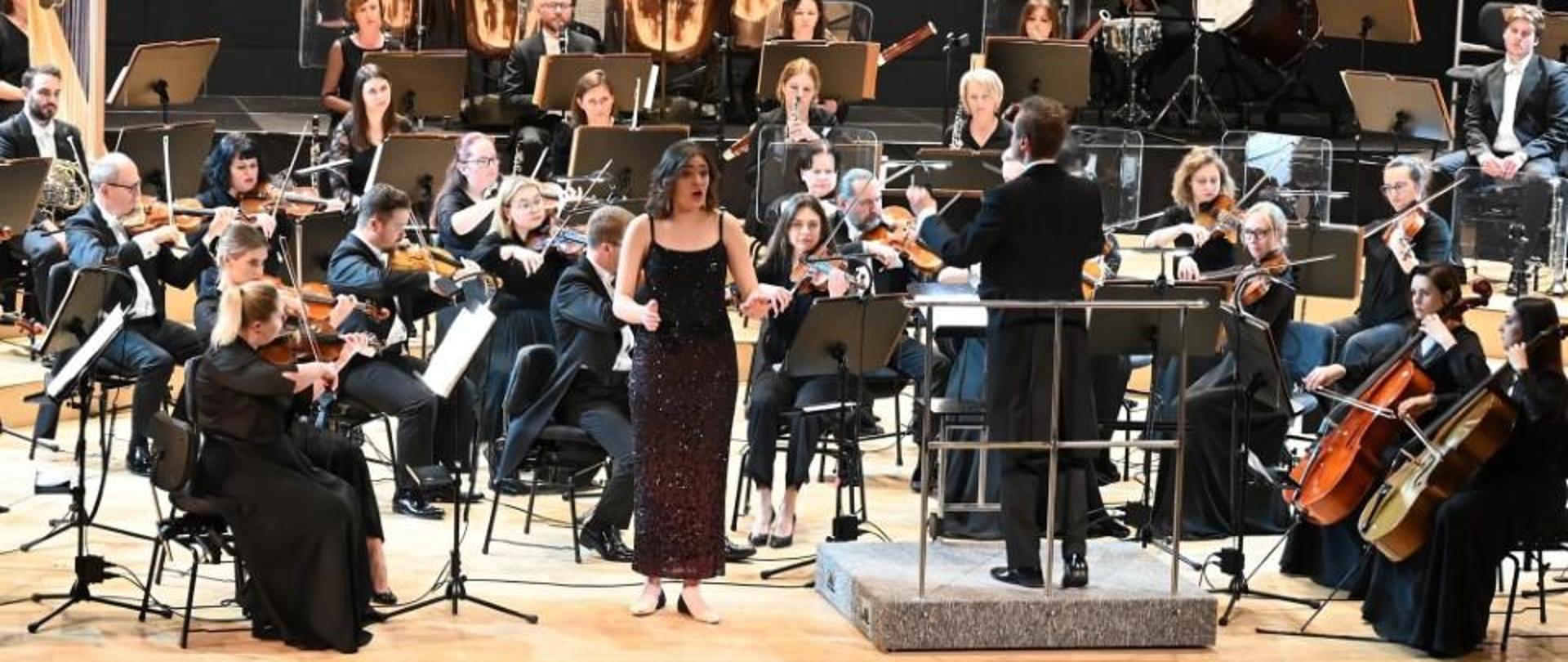 Zdjęcie kolorowe, wokalistka na scenie filharmonii w stroju wieczorowym w otoczeniu orkiestry symfonicznej.