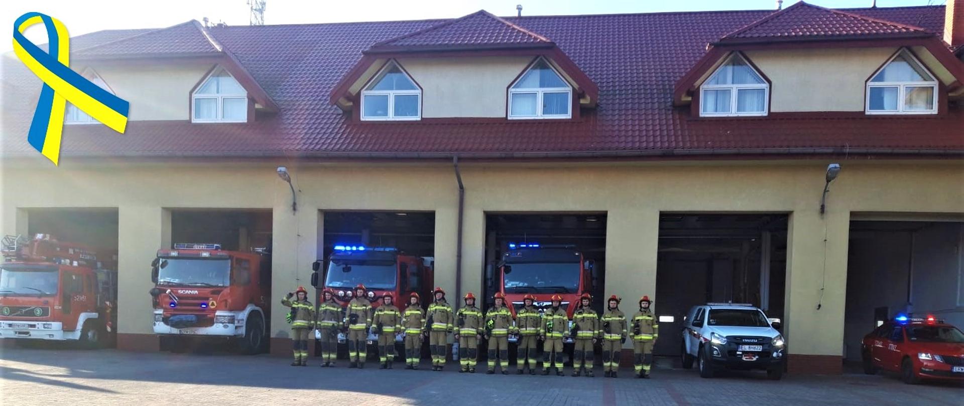 Na zdjęciu, w jednym szeregu stoi 13 strażaków na zbiórce, a za nimi znajdują się samochody strażackie oraz budynek Komendy Powiatowej Państwowej Straży Pożarnej w Rawie Mazowieckiej. Zdjęcie zrobione na wprost. W lewym górnym rogu, grafika wstążki w kolorach żółto-niebieskim.
