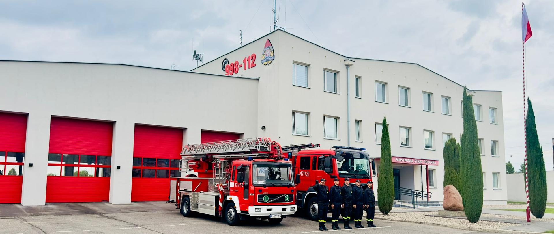 5 strażaków oddaje honory, przed dwoma pojazdami pożarniczymi, w tle budynek straży pożarnej, po prawej powiewająca flaga RP.