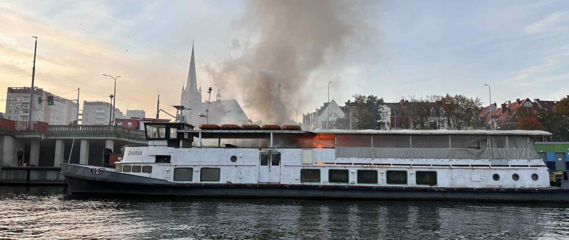 Statek o nazwie „DARIA” stoi zacumowany przy nabrzeżu, z górnej części pokładu wydobywa się intensywny dym i widoczne są płomienie; w tle panorama miasta z kościołem i budynkami mieszkalnymi o zachodzie słońca.