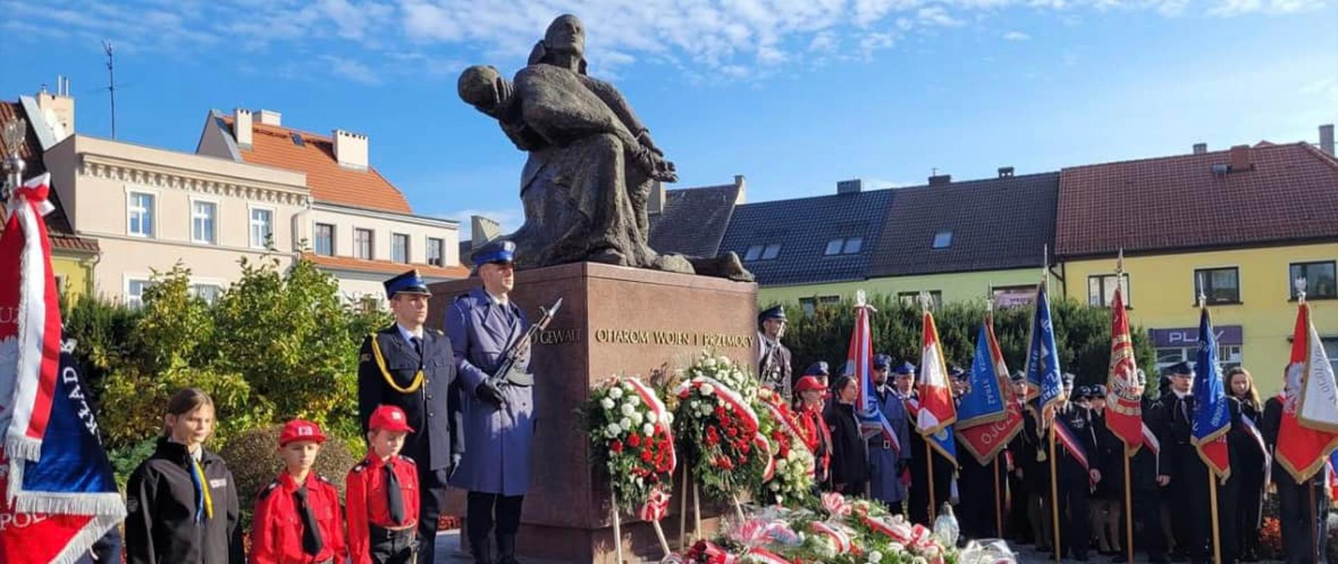Na zdjęciu znajdują się poczty sztandarowe, warta honorowa i inne osoby ustawione przy pomniku, w tle widać budynki