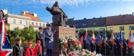 Na zdjęciu znajdują się poczty sztandarowe, warta honorowa i inne osoby ustawione przy pomniku, w tle widać budynki