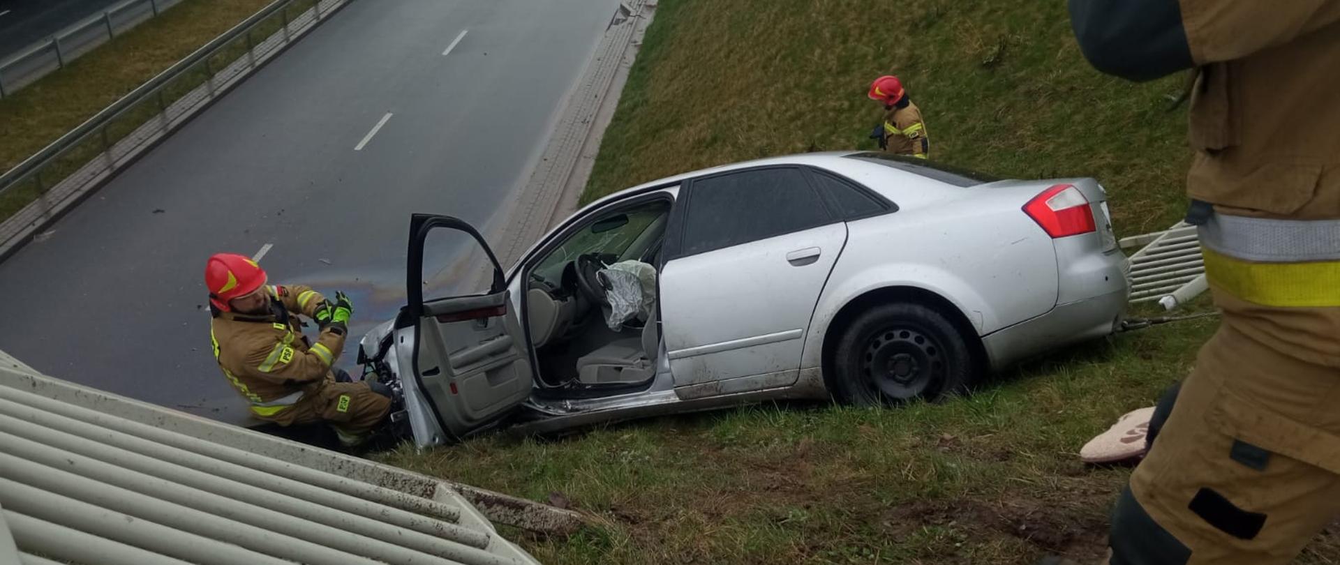 Zdjęcie wykonane w porze dziennej na wiadukcie drogowym w Radomiu na skrzyżowaniu ulic Wojska Polskiego i Lubelskiej. Na zdjęciu widać znajdujący się na nasypie ziemnym pojazd osobowy w srebrnym kolorze. Pojazd uległ zderzeniu z innym pojazdem. Przed pojazdem widać częściowo uszkodzoną barierkę ochronną. Pojazd na skutek uderzenia przerwał barierę staczając się w dół nasypu. Przy pojeździe widać strażaków. Strażacy ubrani w ubrania specjalne w kolorze piaskowym. Na głowach mają założone hełmy ochronne w korze czerwonym. W dole poniżej pojazdu widać jezdnię. 