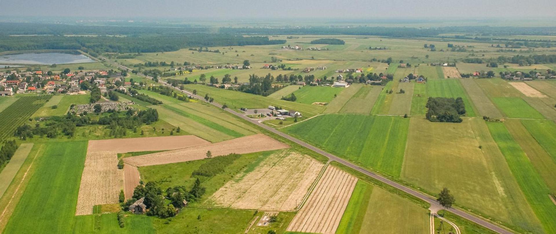 Panorama wiejskiego krajobrazu z lotu ptaka, przedstawiająca rozległe pola uprawne w różnych odcieniach zieleni i żółci, przecięte prostą drogą asfaltową. W tle widoczne są zabudowania wsi, pojedyncze gospodarstwa oraz zbiornik wodny po lewej stronie. Horyzont rozciąga się w dal z lasami i otwartymi przestrzeniami pod lekko zamglonym niebem.