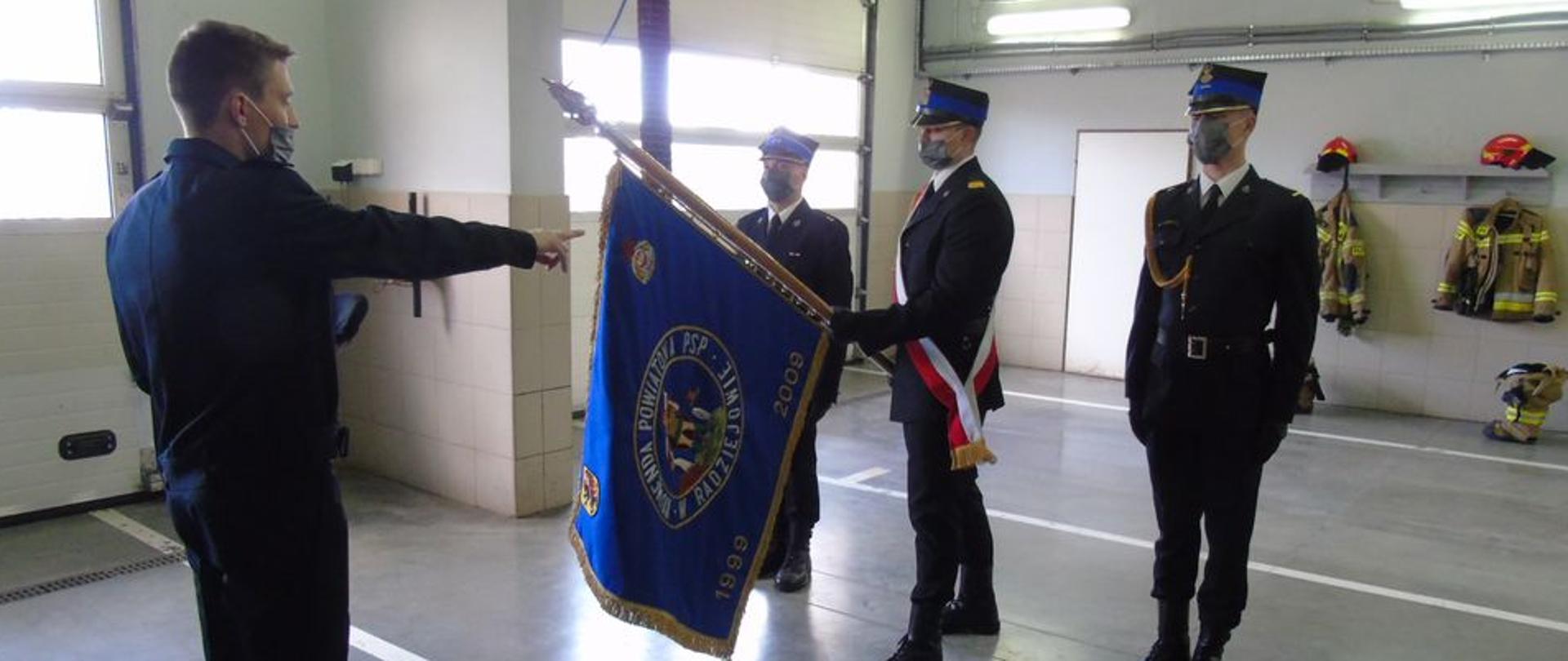 Uroczysta zbiórka w pomieszczeniach garażowych. Na zdjęciu trzech strażaków ubranych w mundury wyjściowe ze sznurem ( gabardyna), na głowach czapki, wszyscy maja maseczki. Jeden ze strażaków trzyma w rękach pochylony do przodu sztandar. Naprzeciwko sztandaru stoi strażak ubrany w mundur służbowy, ma wyciągniętą lewa ręka do przodu, składa ślubowanie.