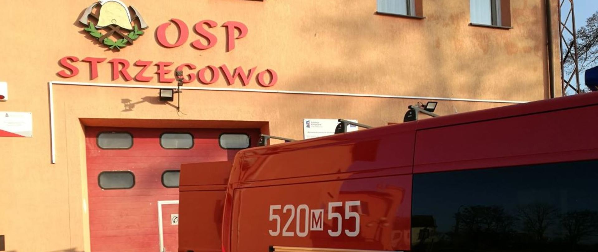 Przed budynkiem remizy OSP Strzegowo stoi zaparkowany pojazd pożarniczy typu BUS, który ma otwarte tylne drzwi. 