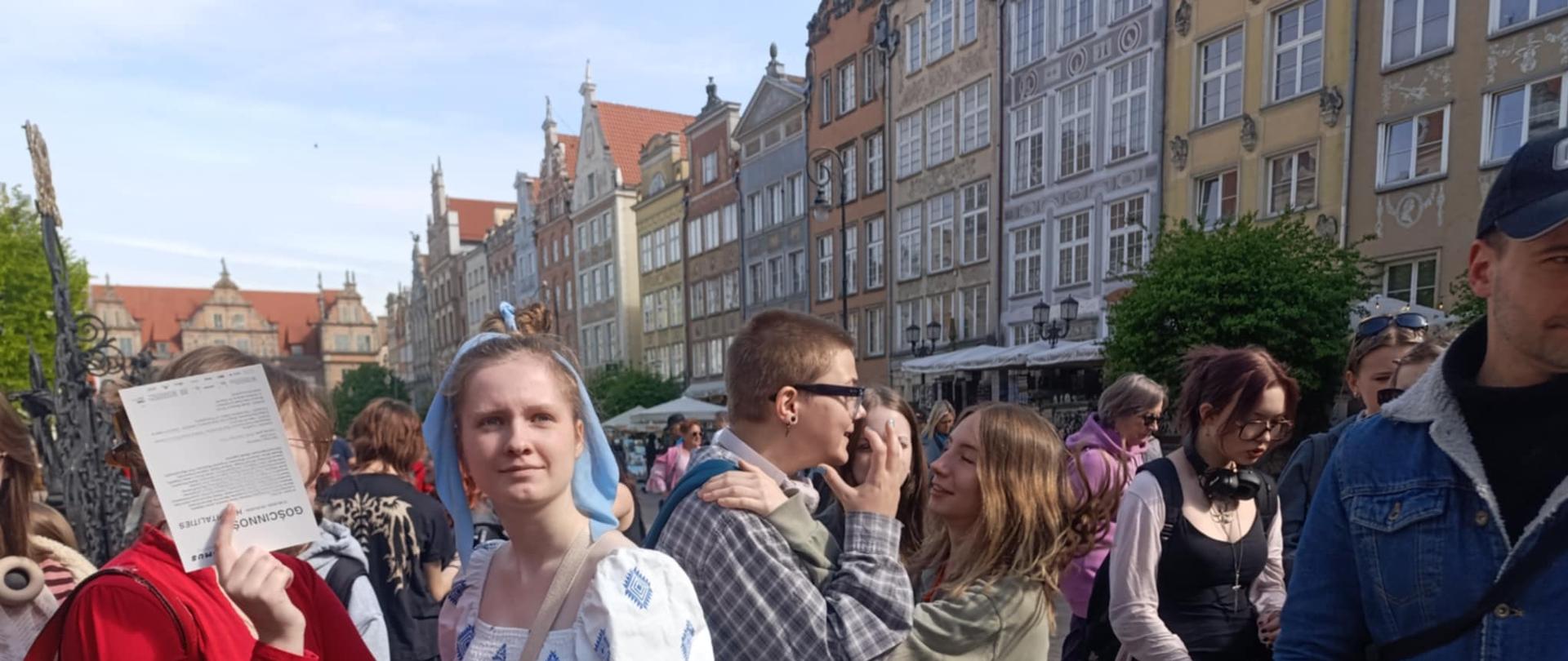 Grupa młodych ludzi stoi na brukowanej ulicy wśród zabytkowych kamieniczek w centrum Gdańska. Na pierwszym planie znajduje się dziewczyna w białej sukni z niebieskim wzorem i niebieską kokardą we włosach, obok niej osoba w czerwonej bluzie zasłania twarz kartką papieru. W tle widać inne osoby, w tym młodzież rozmawiającą ze sobą i turystów, a także charakterystyczną zabudowę gdańskiego Starego Miasta z ozdobnymi fasadami kamienic. Panuje słoneczna pogoda, a atmosfera na zdjęciu jest radosna i towarzyska.