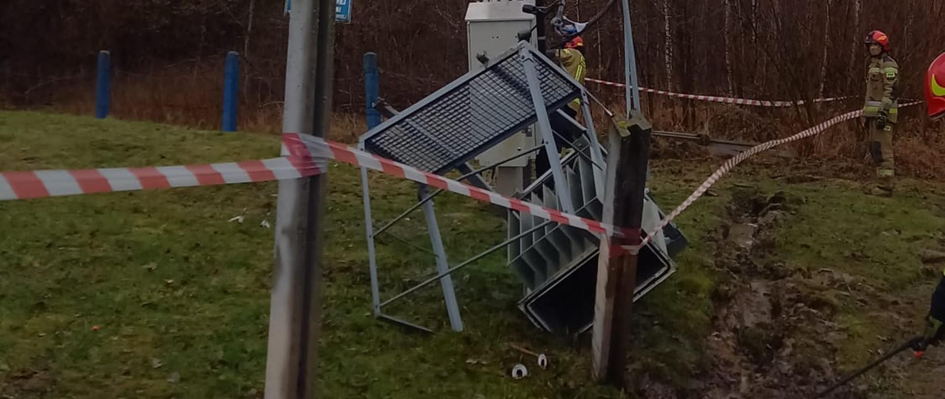 Dzień. Słup energetyczny (betonowy) linii średniego napięcia. U podstawy słupa znajduje się transformator umieszczony w jasnej obudowie. Część przewodów egzegetycznych od transformatora do szczytu słupa energetycznego jest odcięta, zwisa. przed słupem widać leżące na ziemie metalowe elementy konstrukcji transformatora. Z transformatora wycieka olej do gruntu W tle las. Słup odgrodzony taśmą ostrzegawczą w w czerwono białe ukośne paski. Z prawej strony widać dwóch strażaków w ubraniu specjalnym w kolorze piaskowym i czerwony hełm 