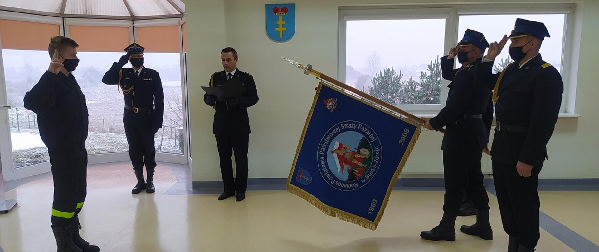 Na zdjęciu po prawej stronie poczet sztandarowy z pochylonym sztandarem oddający honor, z lewej strony zdjęcia przed pocztem stoi strażak składający ślubowanie, na środku zdjęcia komendant powiatowy PSP w Busku-Zdroju przyjmujący rotę ślubowania oraz dowódca uroczystości oddający honor. 