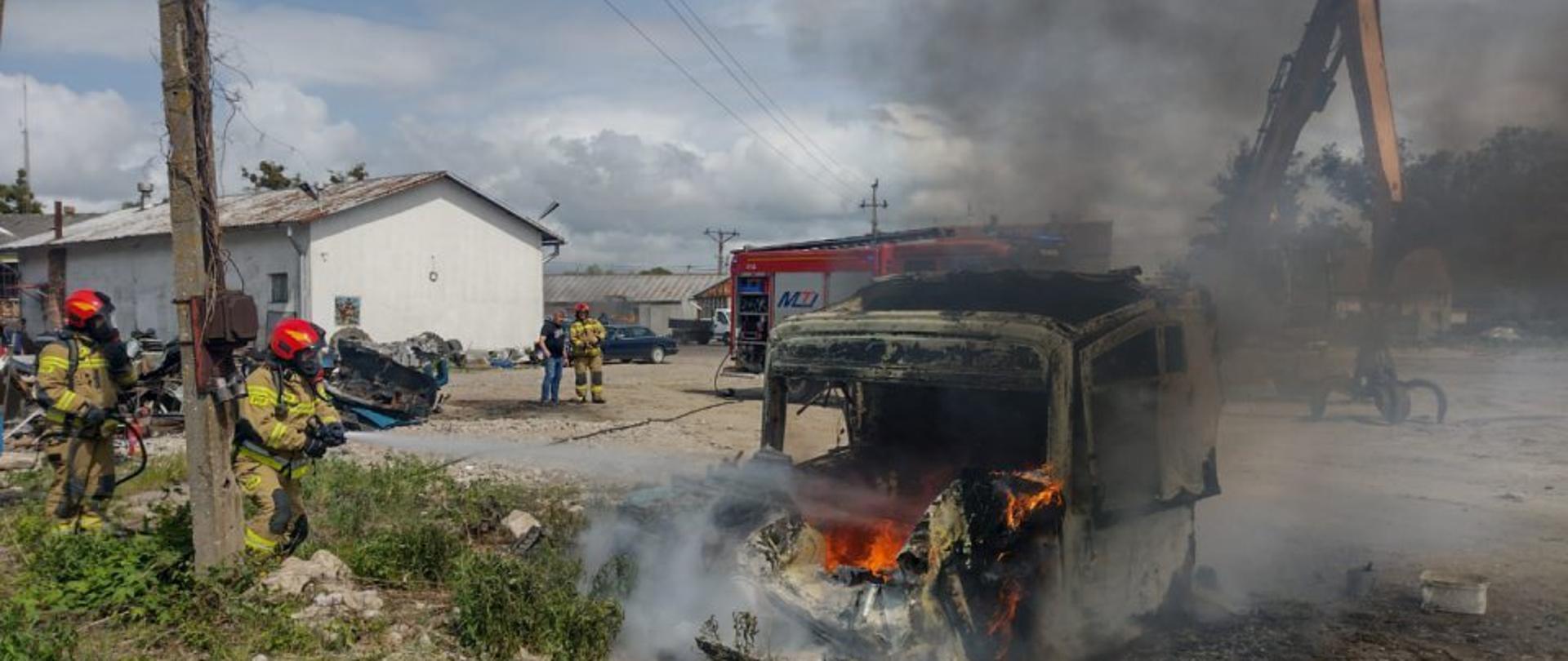 Na placu pali się kabina samochodu ciężarowego, widać płomienie. Z lewej stoi dwóch strażaków w mundurach bojowych i czerwonych hełmach. W tle z tyłu samochód strażacki i budynek. Chmury na niebieskim niebie.