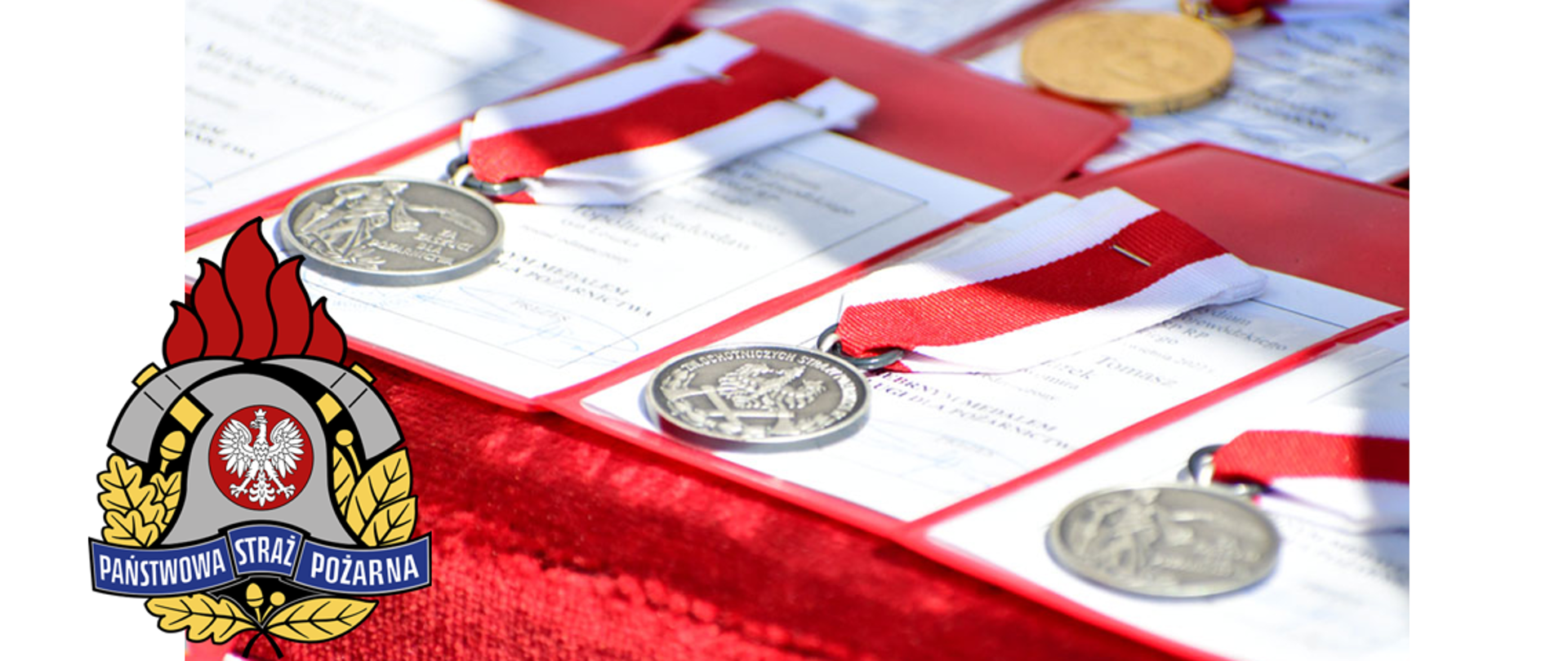 na czerwonym podszyciu widoczne złote i srebrne medale na okoliczność z okazji Powiatowych Obchodów Dnia Strażaka. W lewym dolnym rogu zamieszczone logo Państwowej Straży Pożarnej.
