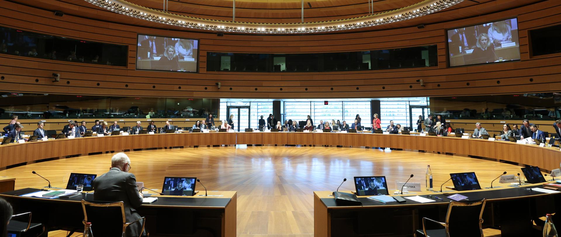 A wide view of the Council of the European Union meeting room, with delegates seated around a large circular conference table. The interior features wooden wall panels and ceiling, as well as a large oval lighting installation. Screens display footage from the meeting, while microphones, monitors and documents are arranged on the tables.