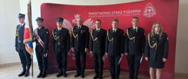 7 strażaków i 1 strażaczka, stoi w mundurach na tle ścianki z logo PSP. Wszyscy ubrani w mundury wyjściowe. Jeden ze strażaków trzyma sztandar. 