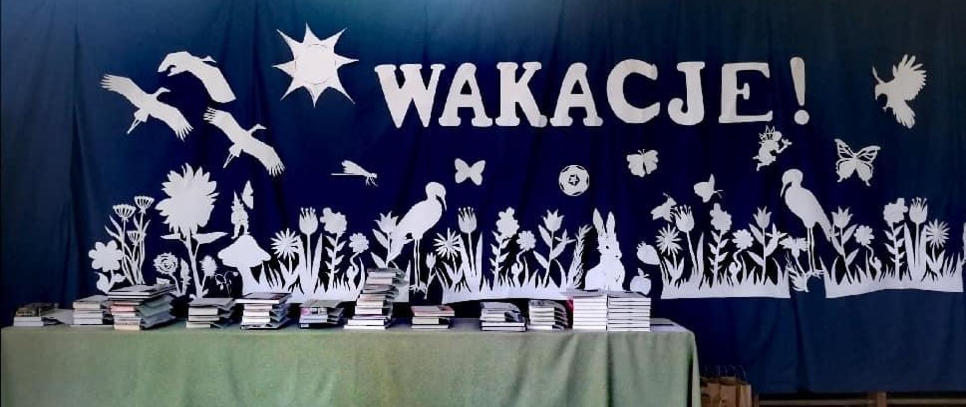 Na zdjęciu stół z nagrodami dla uczniów i świadectwami z wyróżnieniem. W tle dekoracja - na granatowym tle wycięte w białym kartonie kwiaty, ptaki, zwierzęta, które kojarzą się z wakacjami