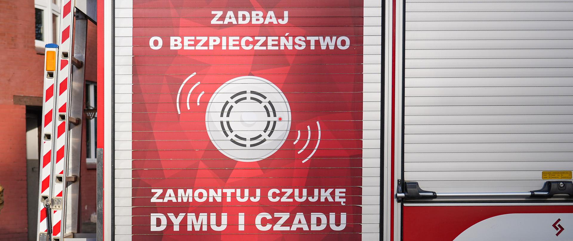 djęcie przedstawia strażaka stojącego obok wozu strażackiego. Strażak ubrany jest w jasnobrązowy mundur i stoi tyłem do otwartej rolety ładunkowej pojazdu. Na bocznej ścianie wozu znajduje się duża czerwona naklejka z białym napisem: „ZADBAJ O BEZPIECZEŃSTWO ZAMONTUJ CZUJKĘ DYMU I CZADU” oraz grafiką przedstawiającą czujnik dymu z falami dźwiękowymi. W tle widoczny jest budynek z czerwonej cegły z garażem.