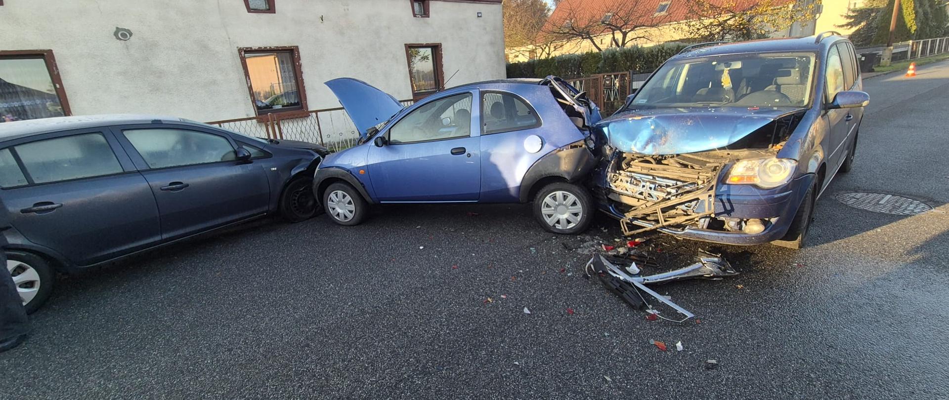 Na zdjęciu znajdują się wraki pojazdów stojące na jezdni, w tle budynek mieszkalny