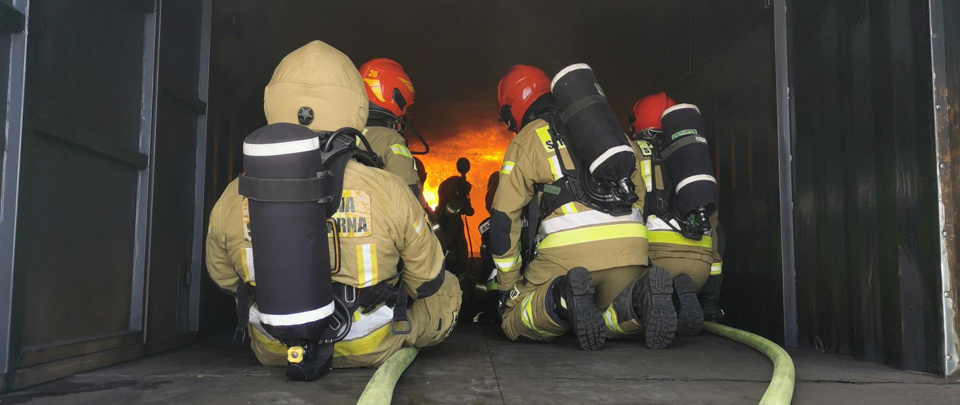 kleczący strażacy w jasnych mundurach bojowych i w hełmach, przed nimi ogień