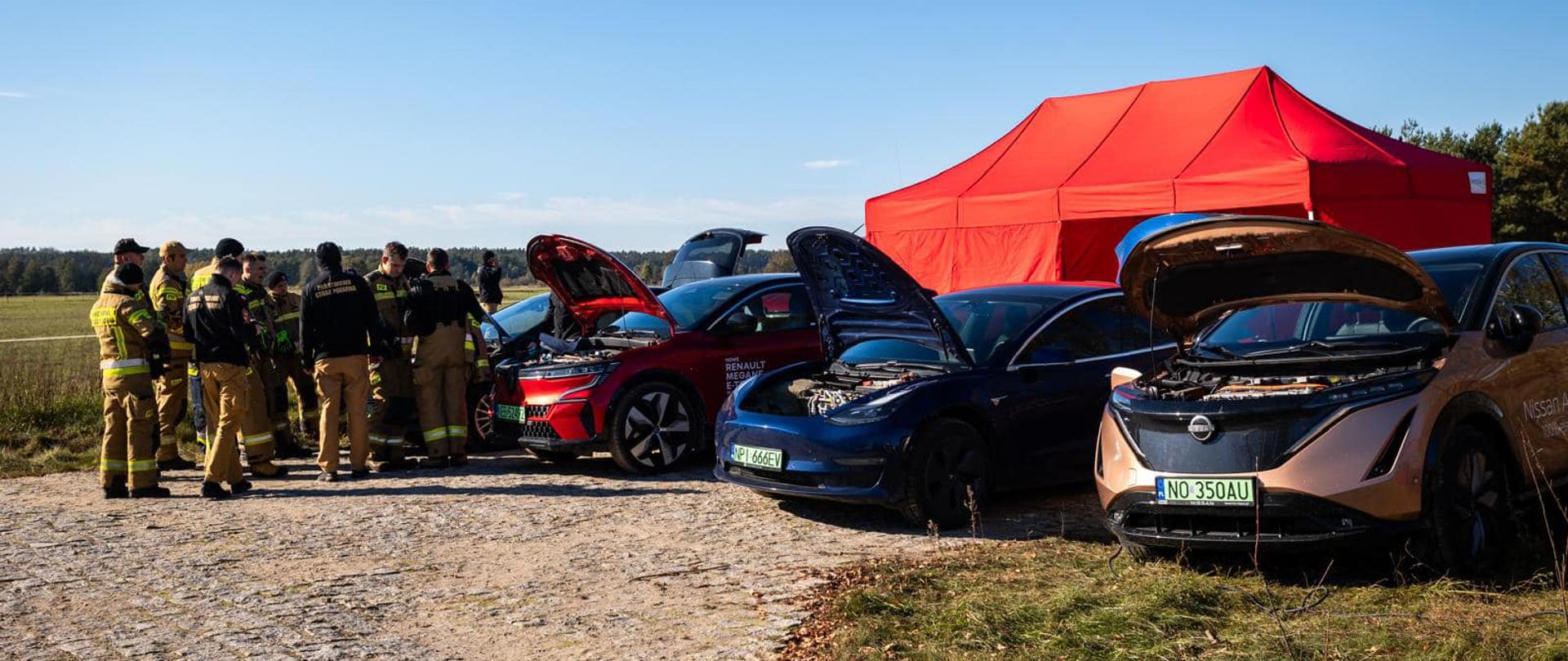 Na zdjęciu znajdują się samochody elektryczne, które stanowiły materiał edukacyjny wykorzystany podczas warsztatów ratowniczych "Ruda 2024". Przy samochodach stoją strażacy - uczestnicy warsztatów.