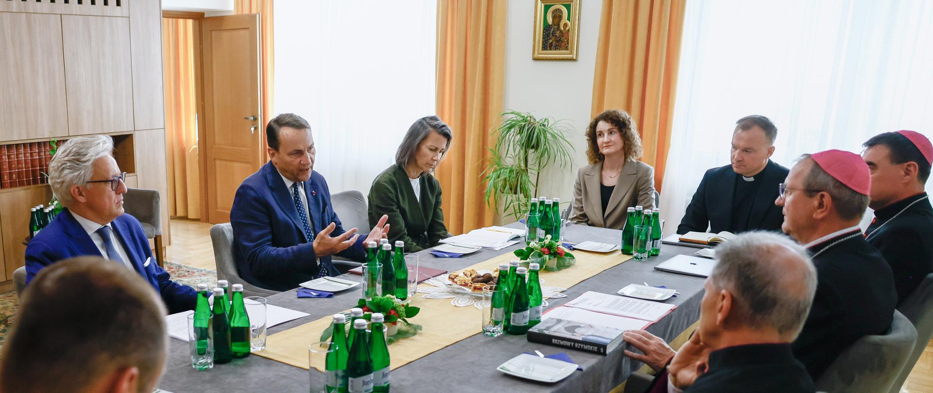 The photo shows a group of people sitting at a table with green water bottles, glasses, and notes on it.
