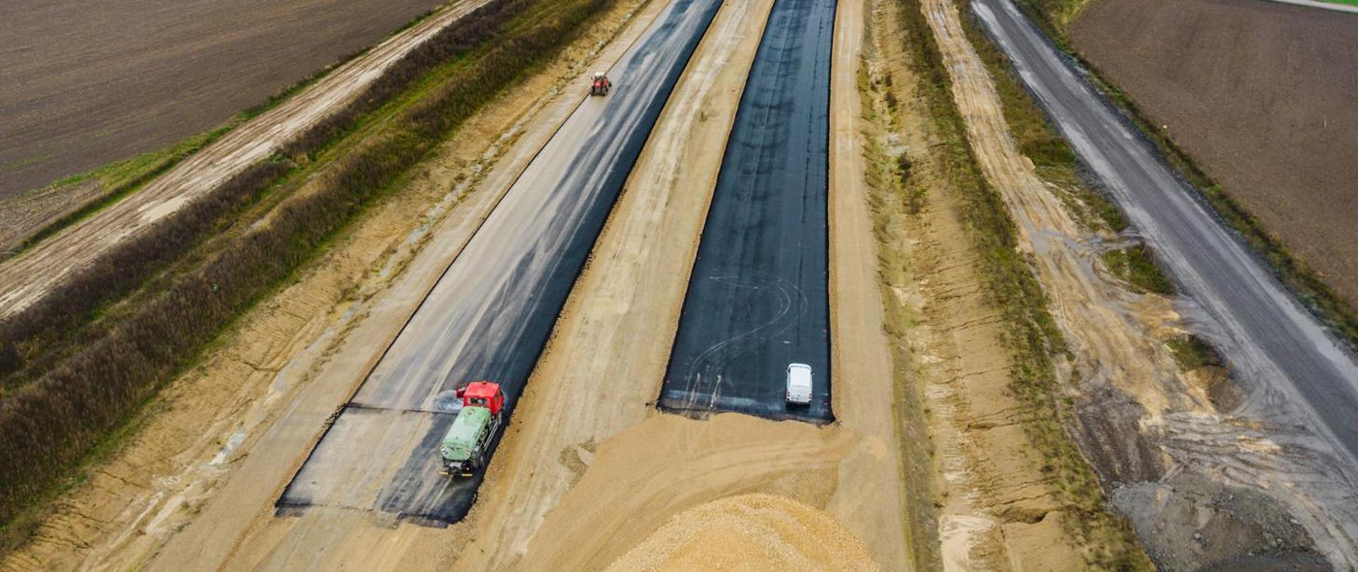 Budowa drogi – widok z góry na dwa pasy asfaltowane w trakcie układania nawierzchni, otoczone ziemnymi nasypami i polami.