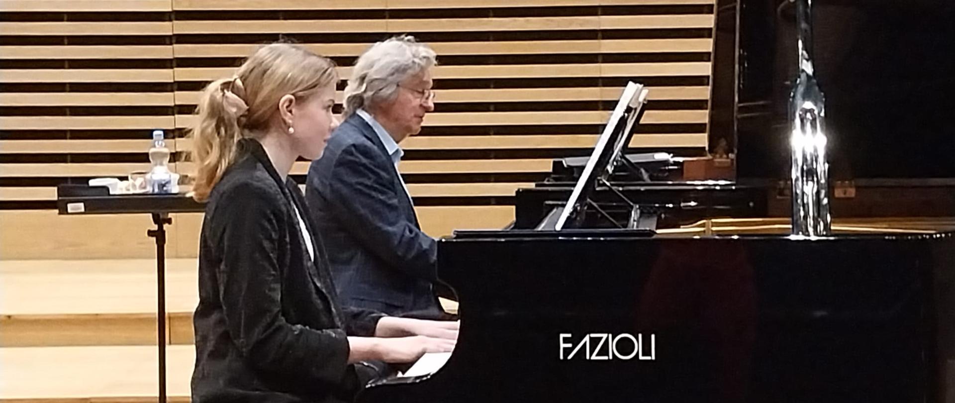 Dziewczyna gra na fortepianie, obok na drugim fortepianie gra mężczyzna.