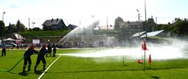 Na zdjęciu widzimy strażaków podających wodę w kierunku pachołków oraz tarczy.
