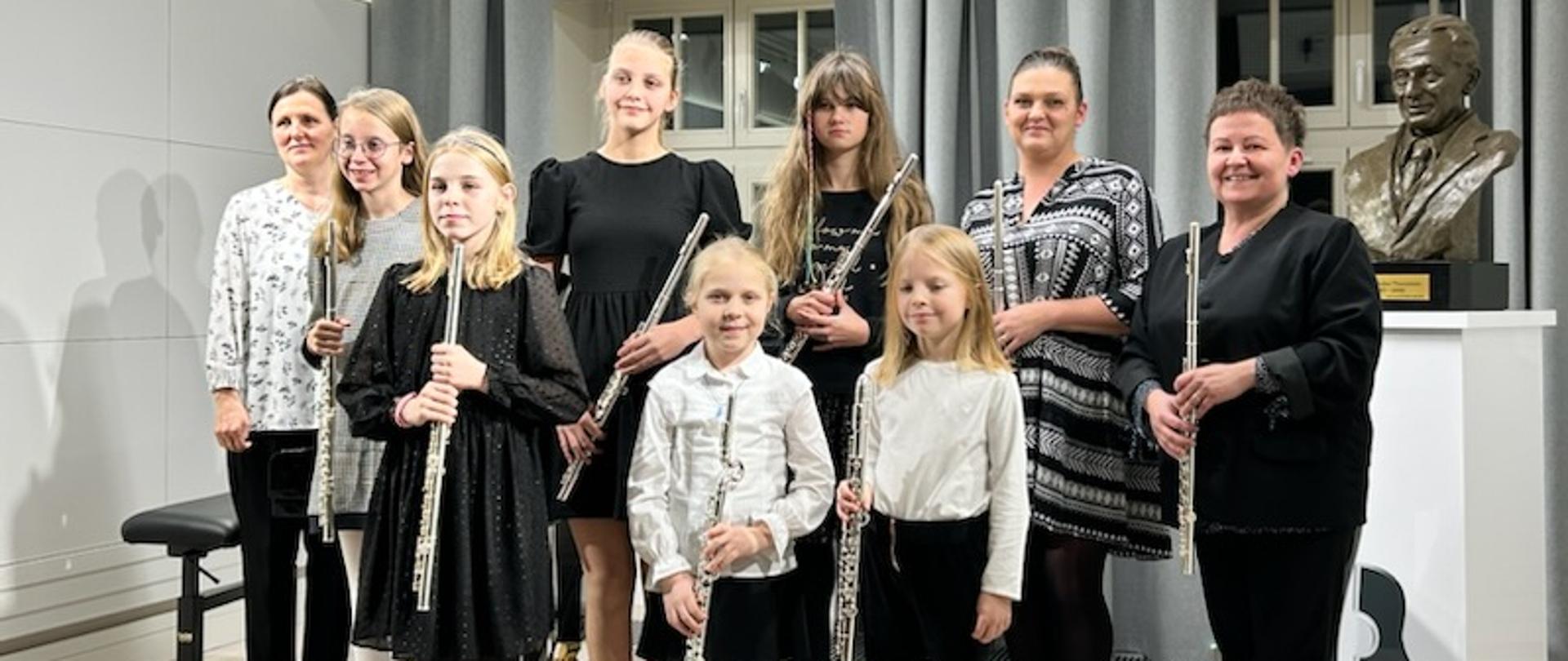 Uczennice ubrane na galowo stoją w dwóch rzędach na scenie i trzymają w dłoniach flety. Z lewe strony stoi pani Edyta Szymańska, a z prawej pani Wioletta Buraczewska. Z tyłu widać odsłonięte okna, z prawej popiersie patrona szkoły na postumencie. 