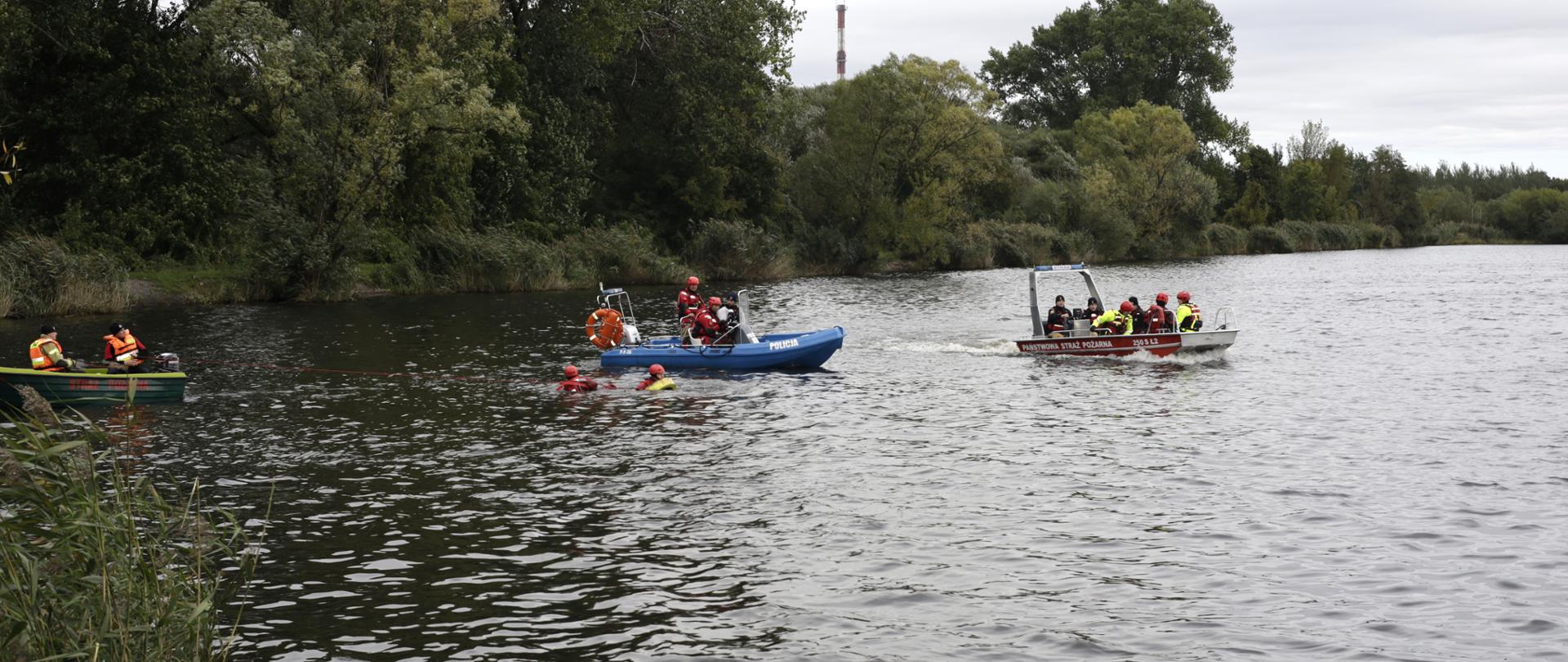 Na zdjęciu widać ćwiczenia ratownicze na zbiorniku wodnym. Na wodzie znajdują się trzy łodzie – zielona, niebieska z napisem „POLICJA” i czerwona z napisem „PAŃSTWOWA STRAŻ POŻARNA”. W wodzie pływa kilku ratowników w kaskach i kamizelkach asekuracyjnych. Akcja odbywa się blisko brzegu porośniętego trzciną i drzewami. Pogoda jest pochmurna, a atmosfera wskazuje na wspólne działania służb ratowniczych.