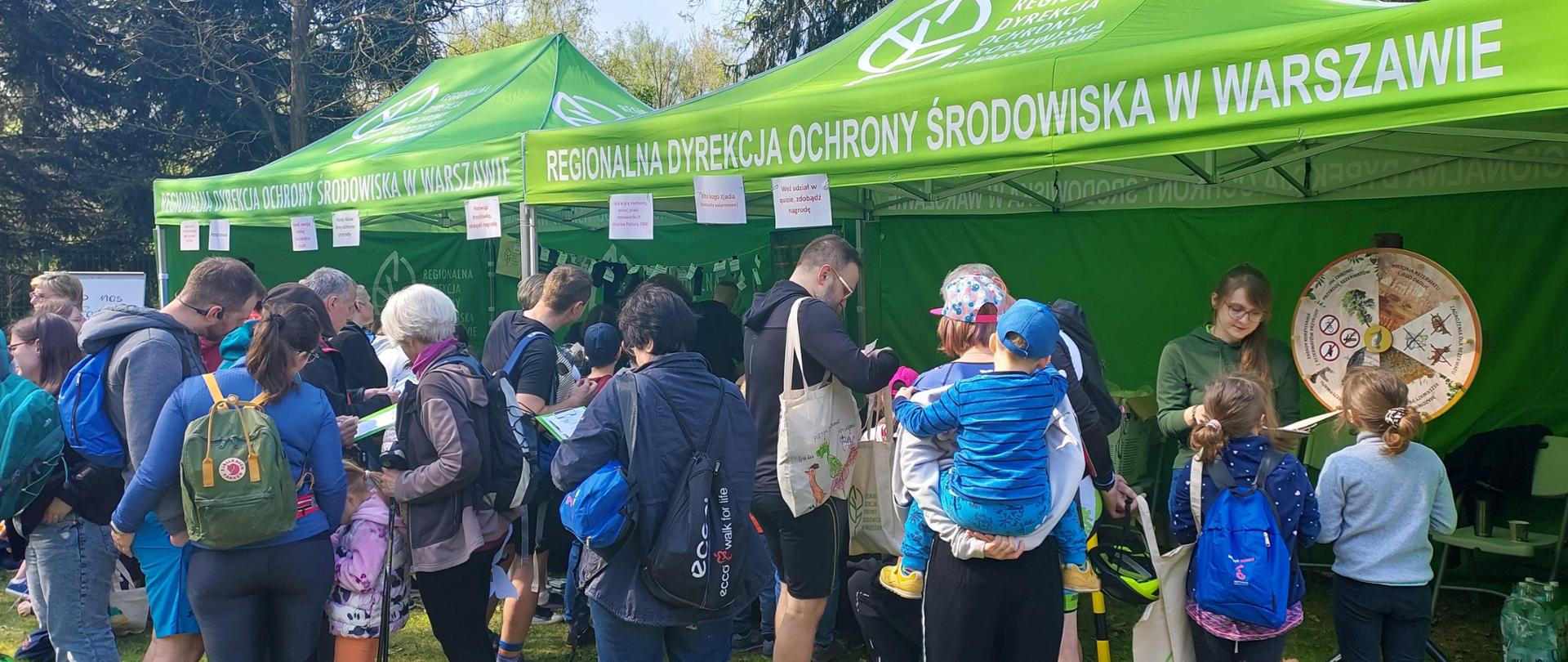 Grupa osób stojących przed zielonymi namiotami, biorących udział w grach edukacyjnych