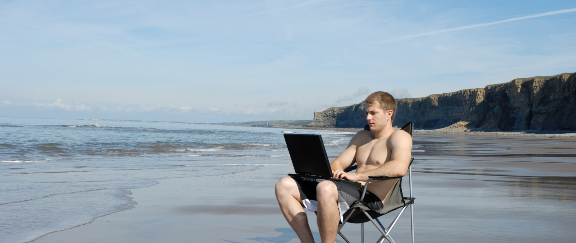 Mężczyzna z laptopem siedzi na krzesełku turystycznym na plaży morskiej, w tle klif.