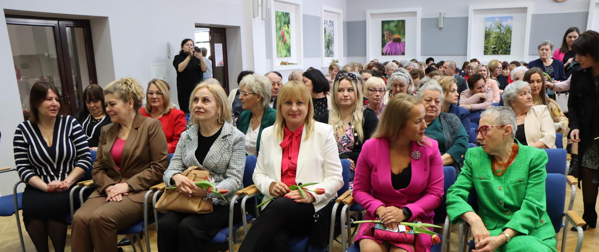 Grupa kobiet siedząca w jasnym pomieszczeniu. Na ścianach wiszą kolorowe obrazy w białych ramkach. Kobiety są ubrane elegancko i kolorowo. Trzymają w dłoniach tulipana. Kobieta w tle robi zdjęcia sali aparatem. 