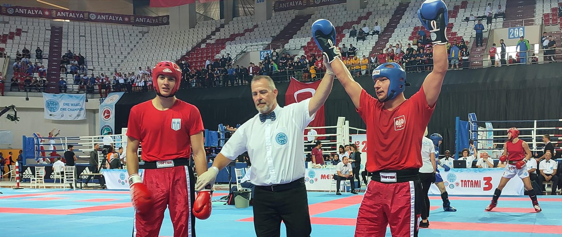 Dwaj zawodnicy stoją na macie po stoczonym pojedynku. Po środku stoi sędzia i unosi ręce zawodnika po prawej stronie w geście zwycięstwa. W tle widać kibiców na trybunach.